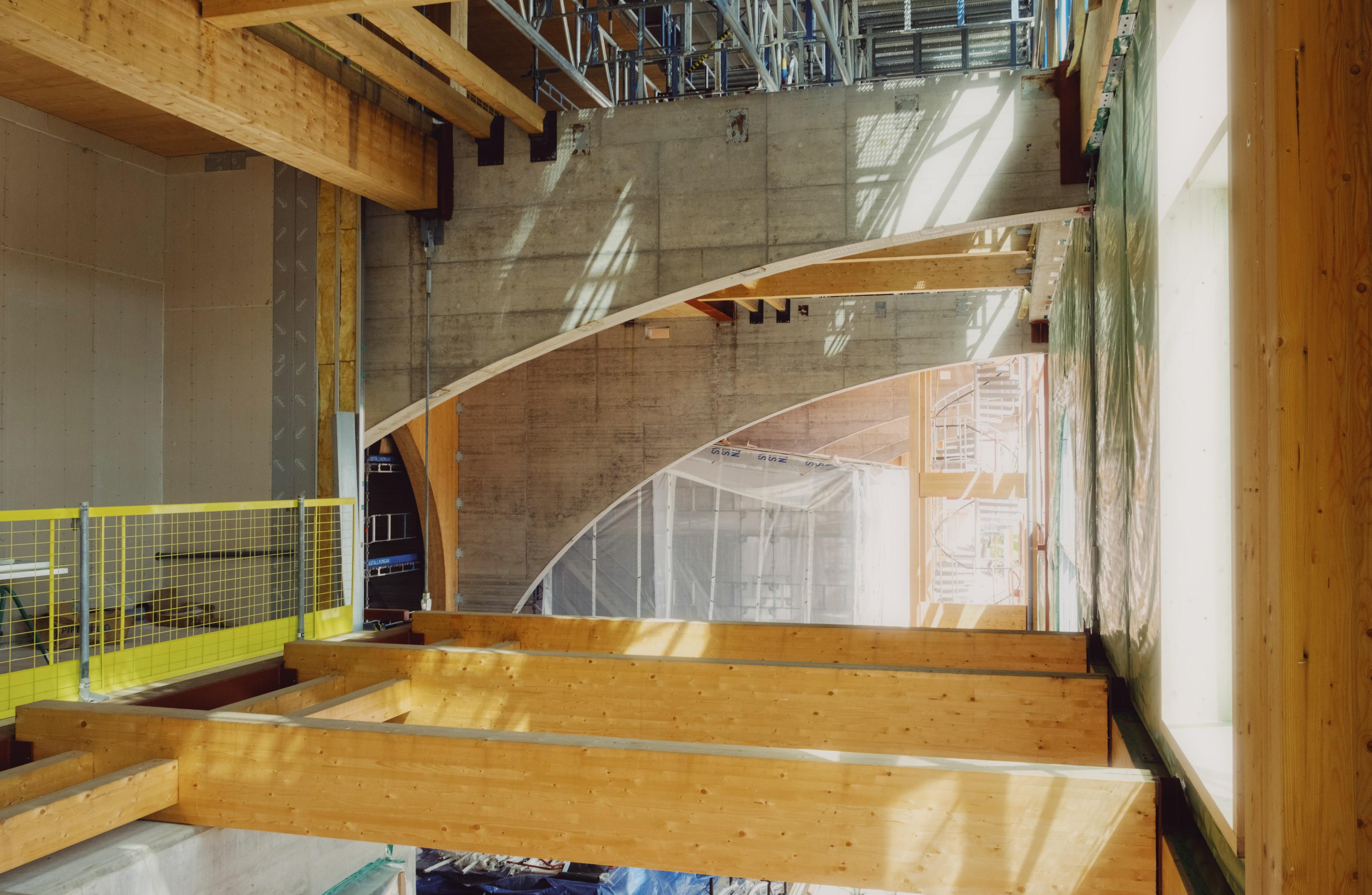 Byggarbetsplats med stora böjda träbalkar och bågar under montering, synligt solljus genom fönster och betongväggar i bakgrunden