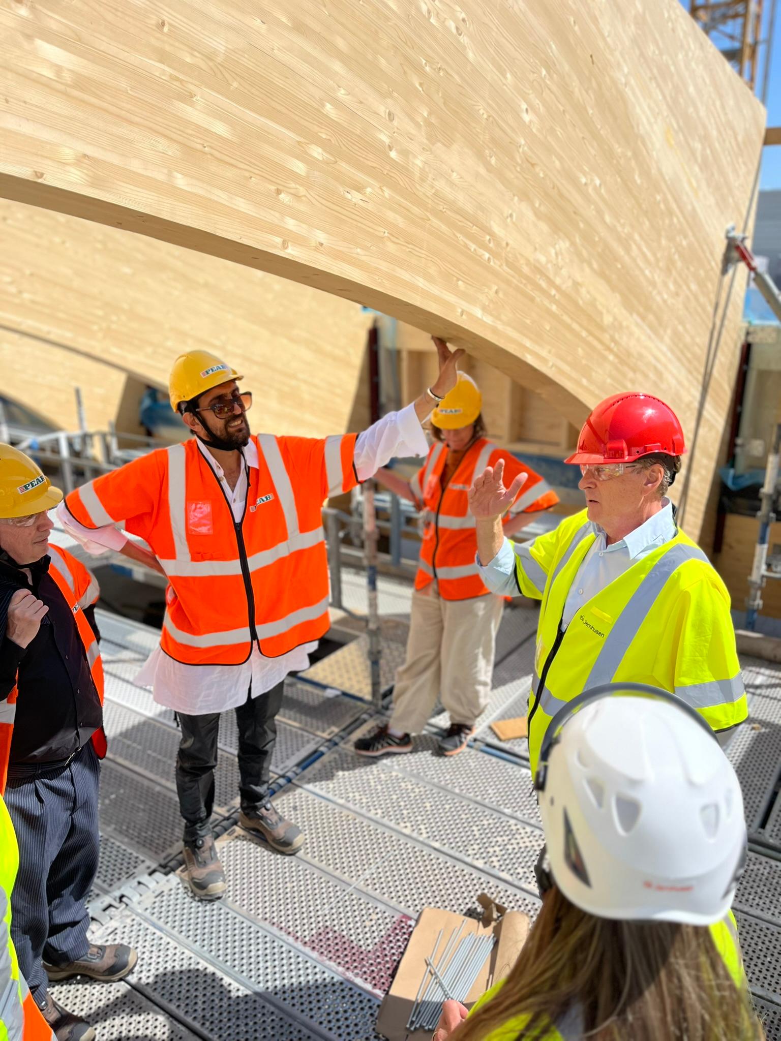 Grupp med byggarbetare och besökare i säkerhetsvästar och hjälmar står under de stora träbågarna på byggarbetsplatsen och diskuterar projektet