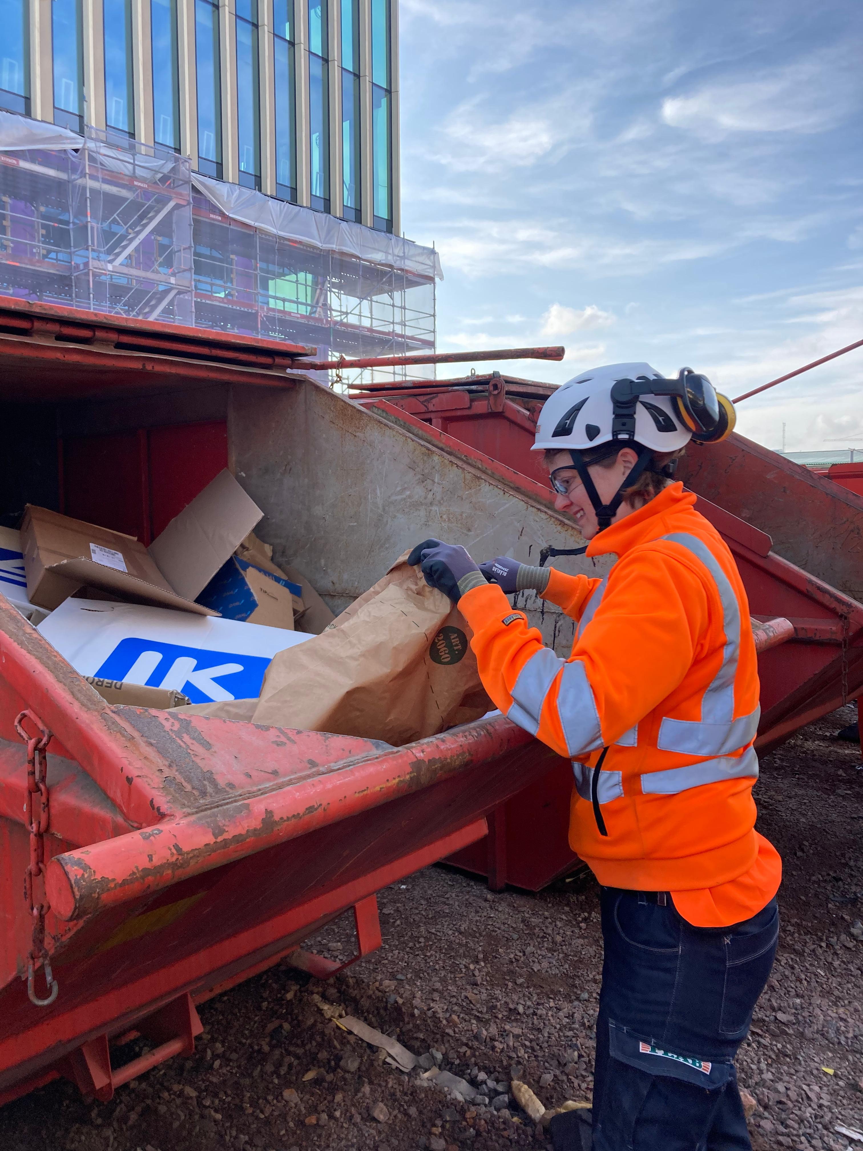 Byggarbetare i orange säkerhetsväst sorterar noggrant material i avfallscontainer.