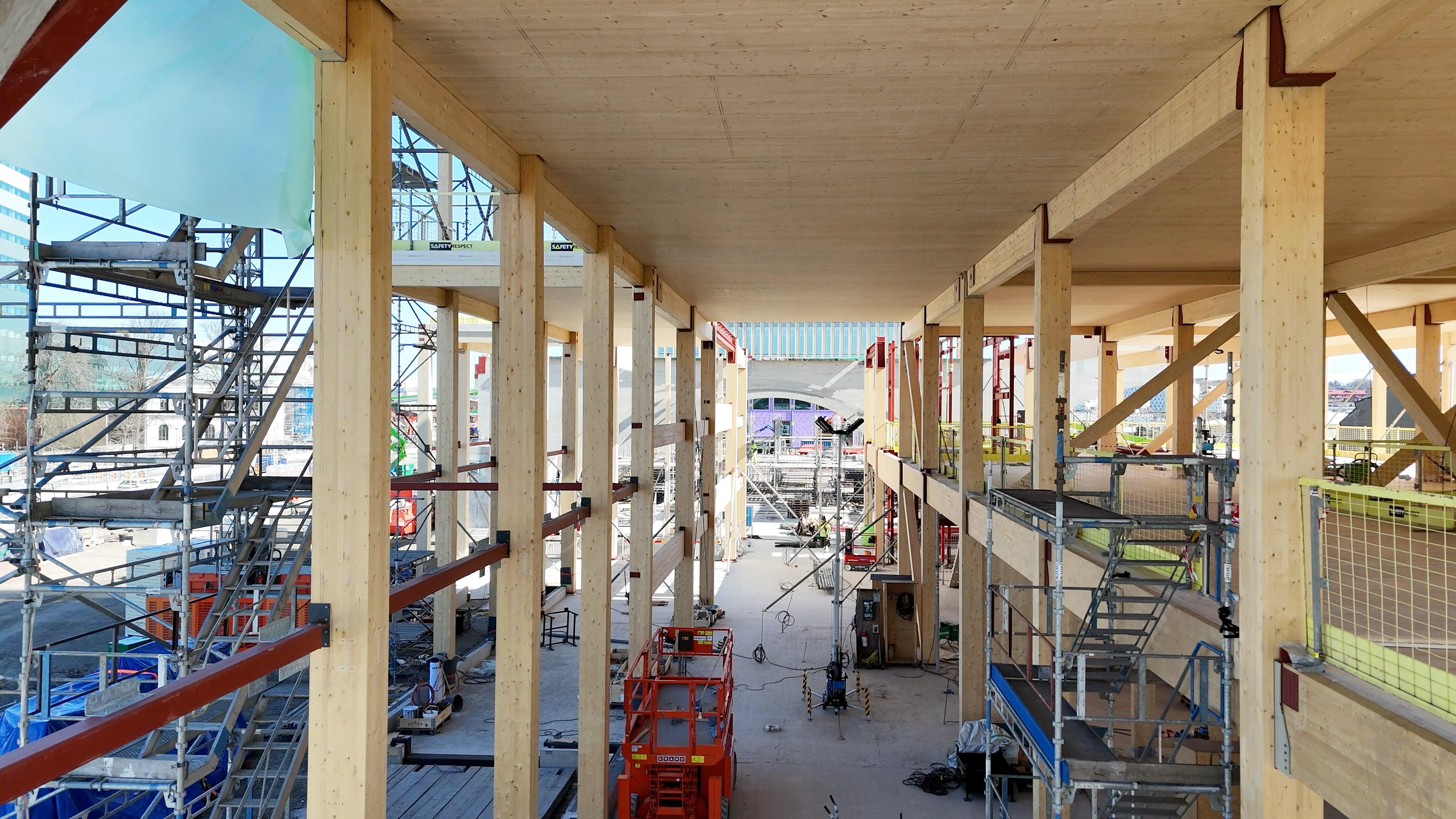 Byggarbetsplats inomhus med massiva träkonstruktioner, synliga limträbalkar i tak och väggar, orange lyftplattformar och genomgående utsikt genom byggnaden mot Göteborgs centrum