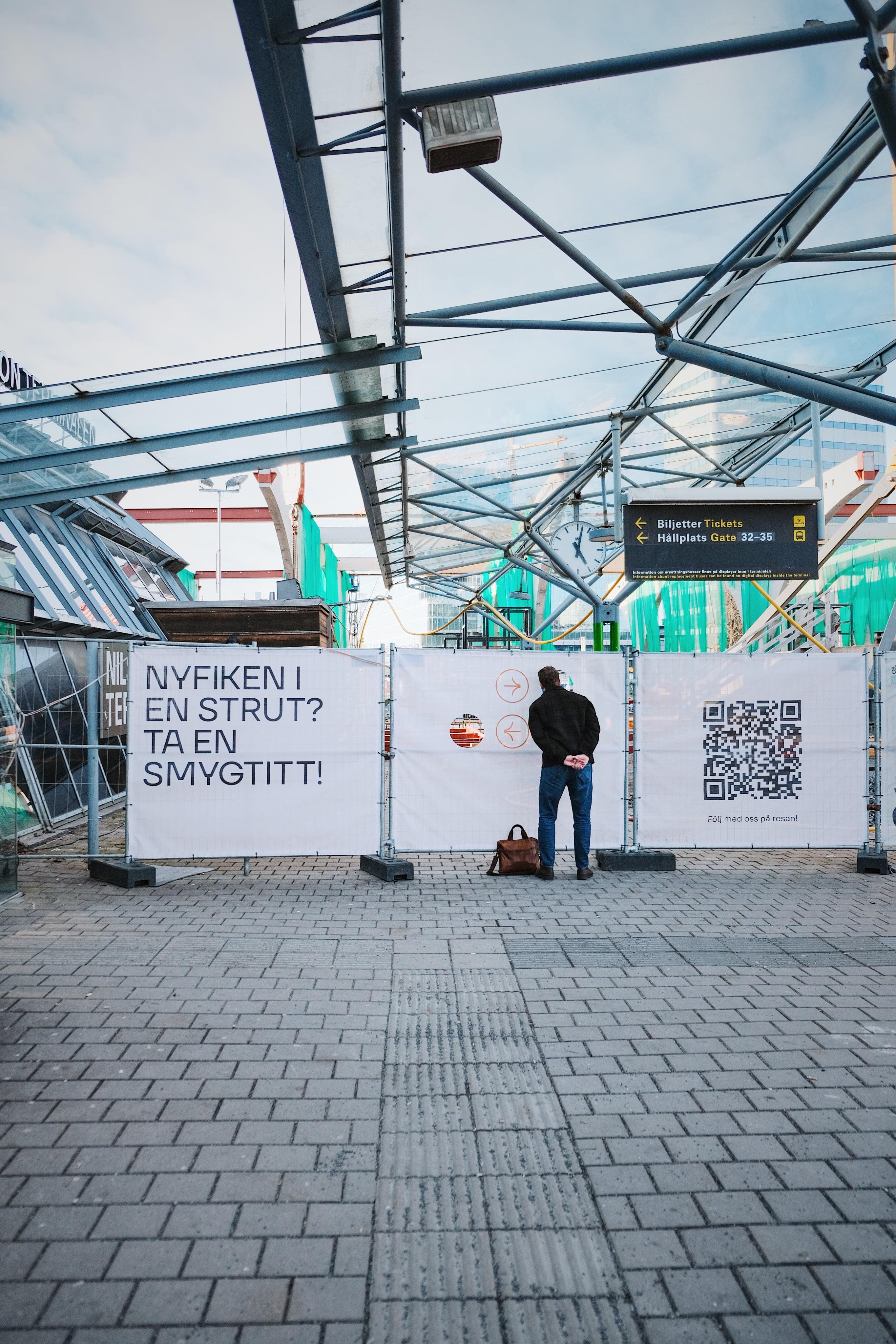 Person vid informationsskärm under glastak med texten 'ÄR DU NYFIKEN PÅ EN STRUT? TA EN SMYGTITT!' och QR-kod.