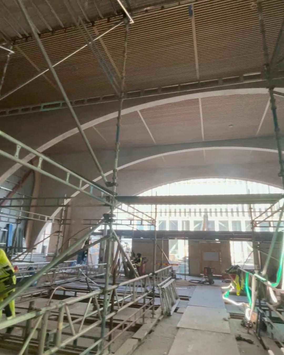 Interiör från pågående byggnation med synlig takstruktur av böjda metallbalkar och träpaneler. Byggställningar, verktyg och byggmaterial ligger utspridda på golvet. I bakgrunden syns stora fönster och ljusa väggar under konstruktion.